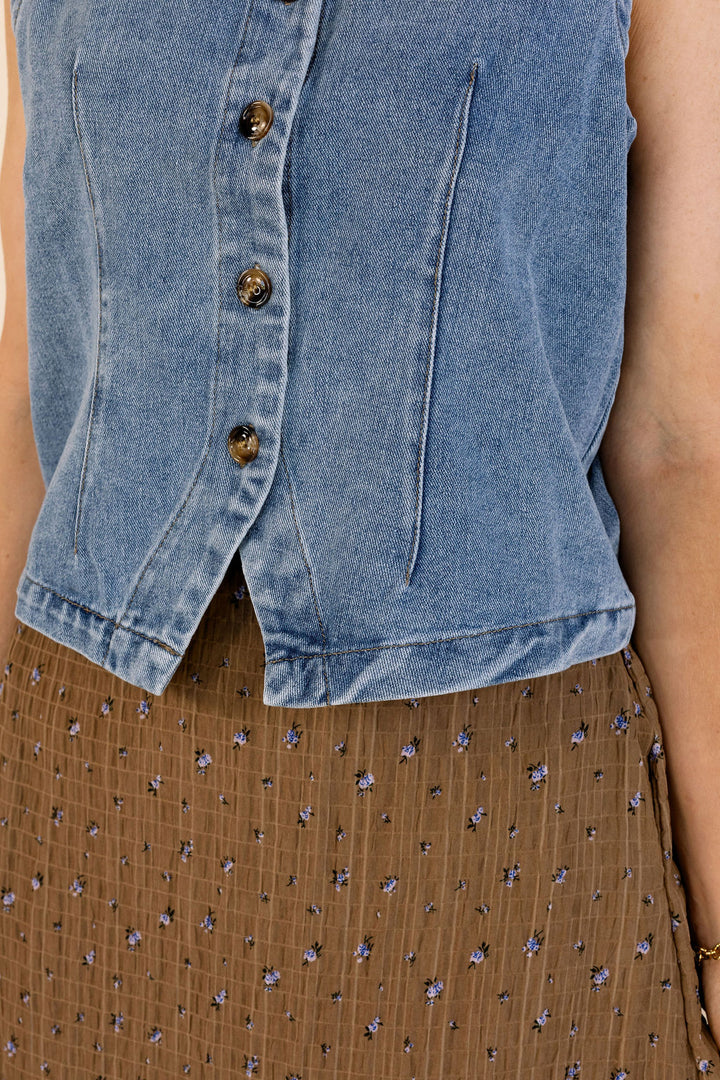 Close-up of a person wearing a light blue denim sleeveless vest with dark buttons over a brown patterned skirt.