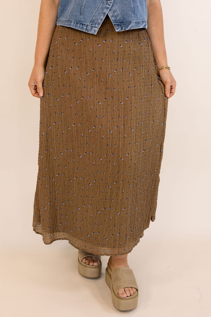 A brown textured midi skirt with a small blue and white floral pattern, worn by a model with a denim top and beige platform sandals against a light beige background.