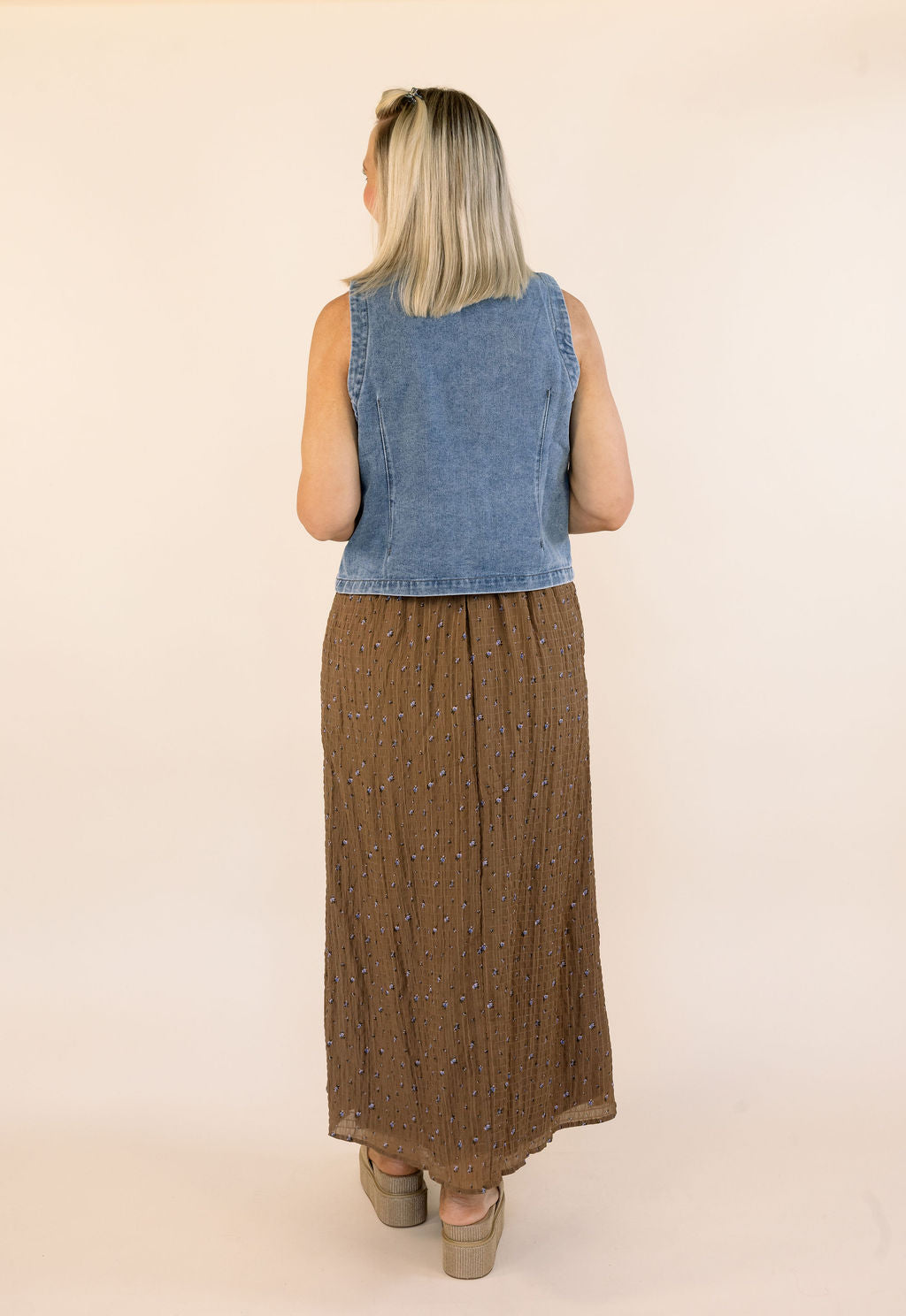 A woman shown from the back wearing a sleeveless blue denim vest and a brown pleated maxi skirt with a subtle speckled pattern, standing against a plain beige studio background.