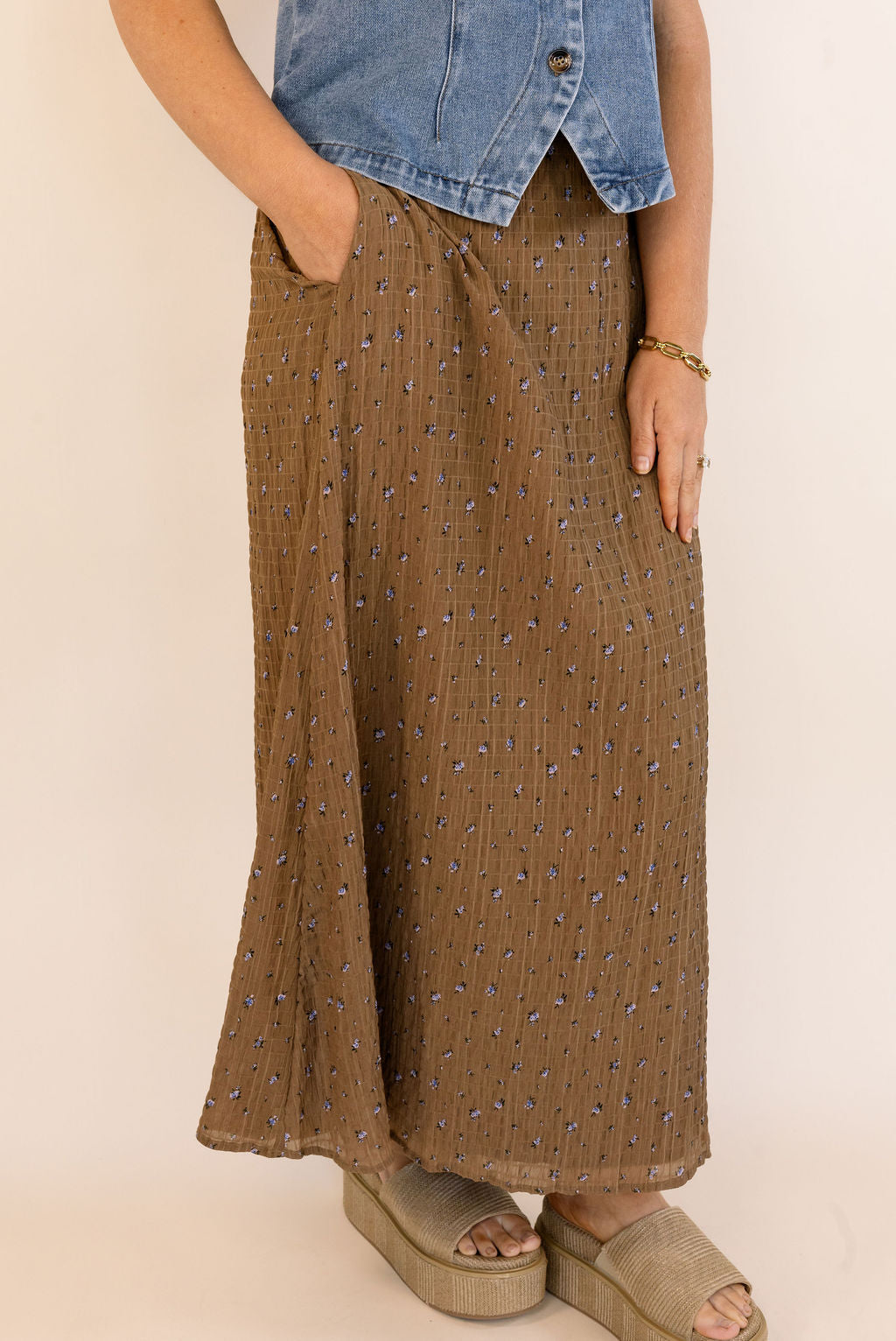 A person wearing a brown maxi skirt with a small blue floral pattern, paired with a denim top and beige platform sandals, shown from the waist down against a plain beige background.