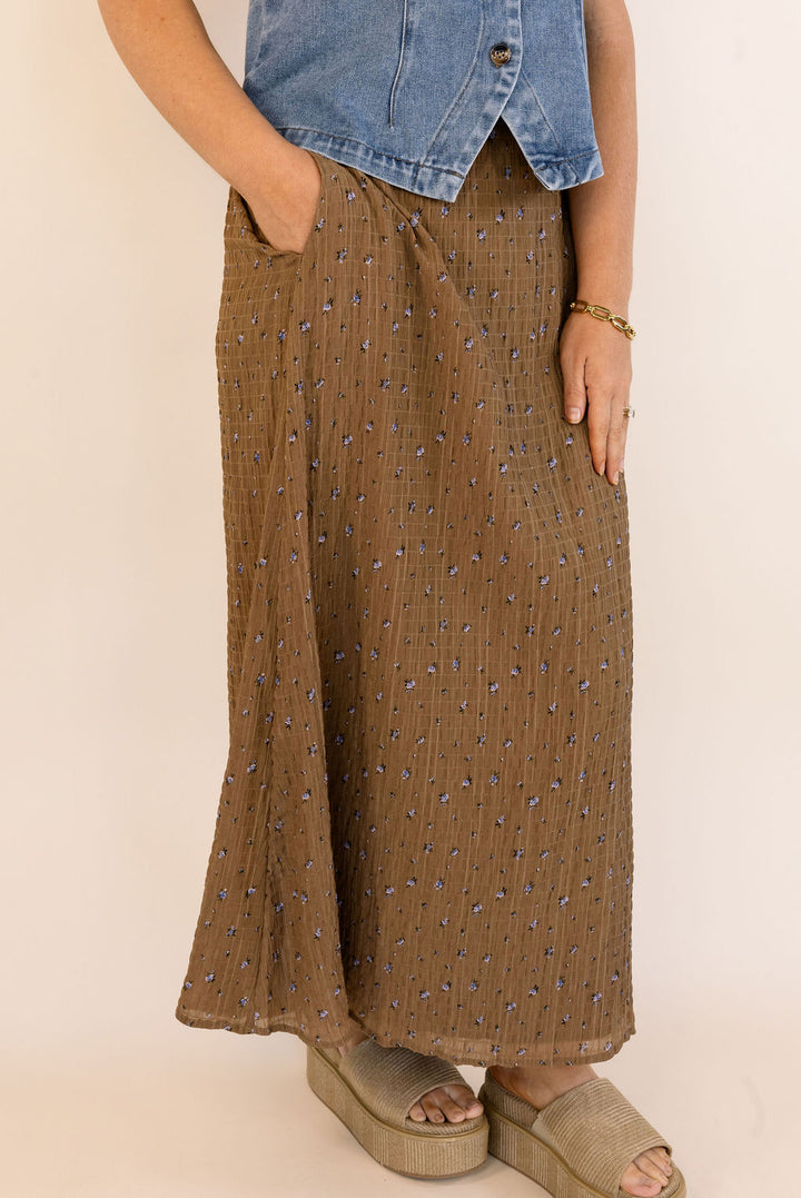 A person wearing a brown maxi skirt with a small blue floral pattern, paired with a denim top and beige platform sandals, shown from the waist down against a plain beige background.
