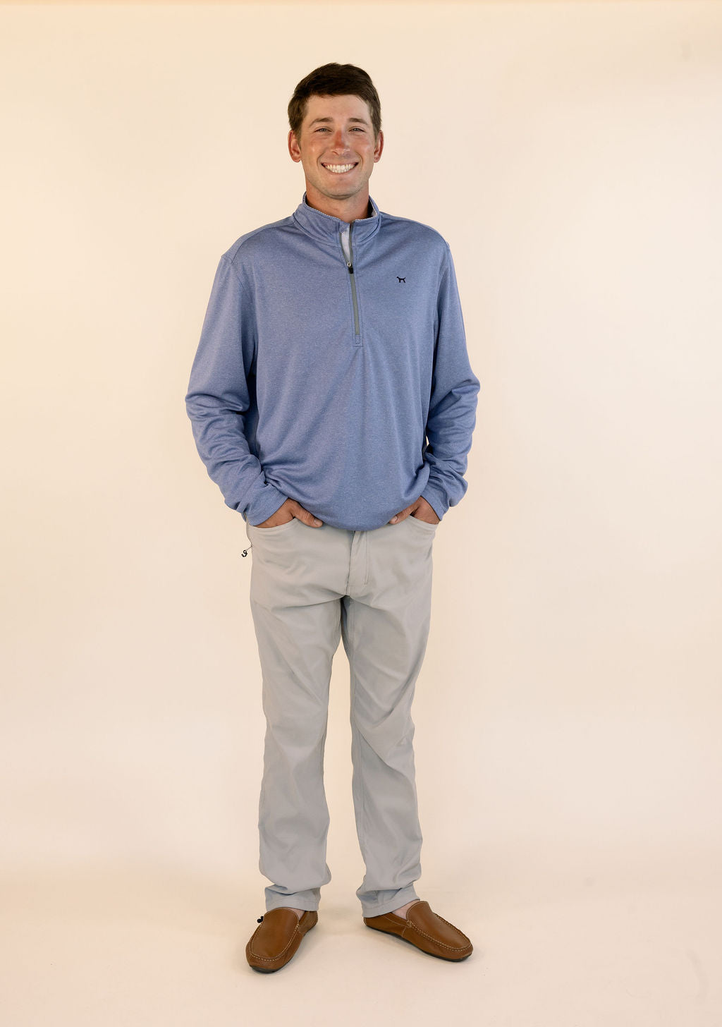 A man wearing a heathered blue quarter-zip pullover, khaki pants, and brown suede loafers, standing against a solid cream-colored studio background.