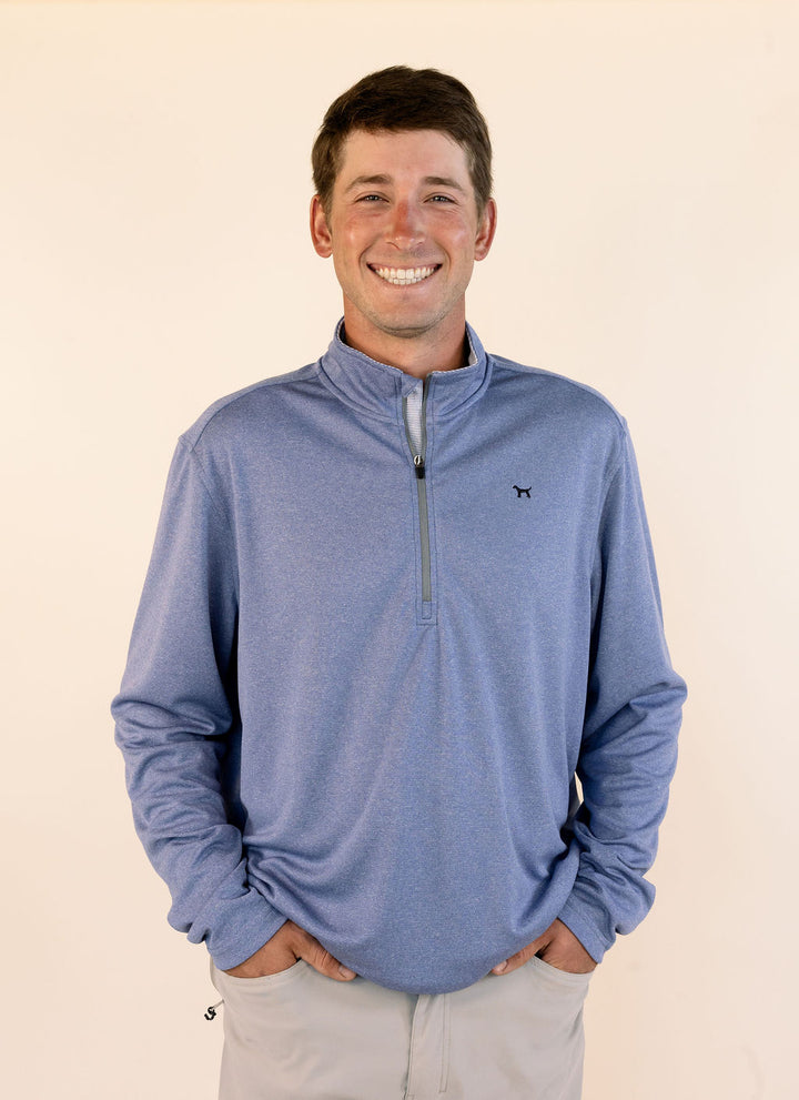 A smiling man wearing a heathered blue quarter-zip pullover sweater with a small black dog logo on the left chest, paired with light gray pants, standing against a solid cream background.