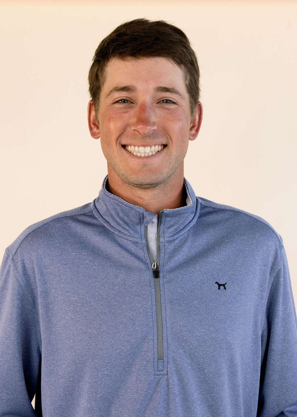 A smiling man wearing a heathered blue quarter-zip pullover with a small black dog logo on the left chest, set against a plain light beige background.