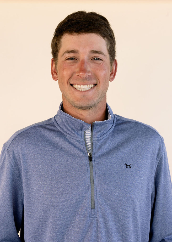 A smiling man wearing a heathered blue quarter-zip pullover with a small black dog logo on the left chest, set against a plain light beige background.