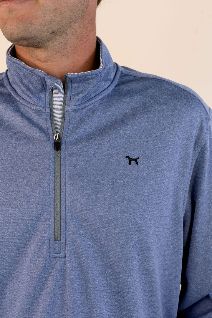 A close-up of a man wearing a heathered blue quarter-zip pullover with a small black dog logo on the left chest, set against a plain beige background.