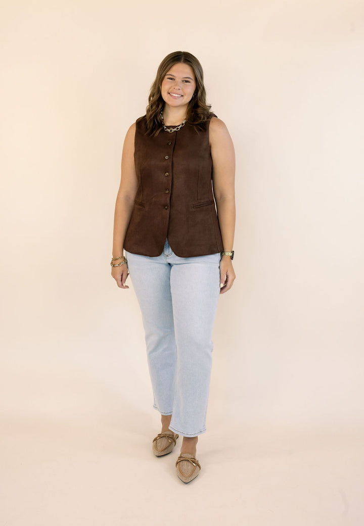 A woman modeling a sleeveless dark brown vest with front buttons, paired with light blue jeans and tan flats, standing against a solid beige studio background.