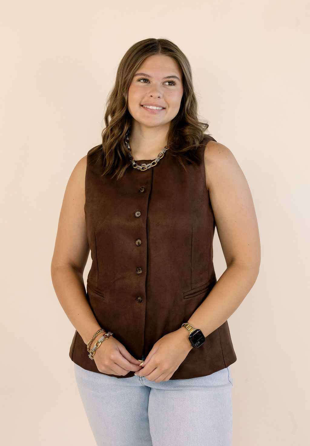 A woman modeling a sleeveless brown vest with a button-down front, paired with light blue jeans, against a solid beige background.