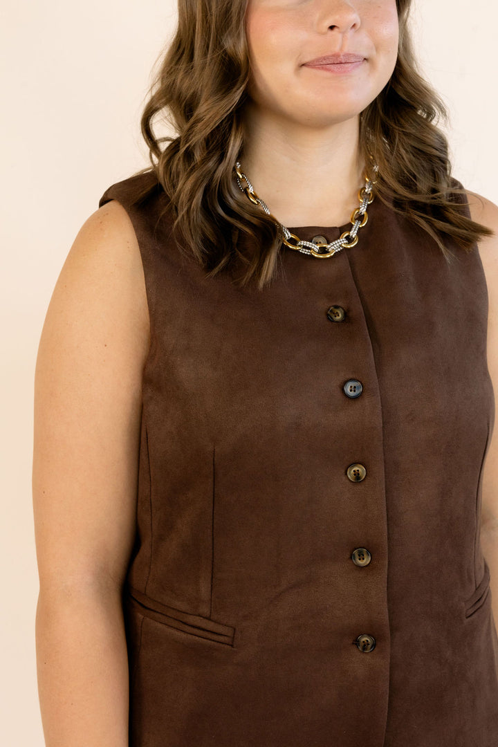 A close-up of a woman wearing a sleeveless brown faux-suede vest with a high collar and a row of decorative buttons down the front, paired with a chunky gold chain necklace.