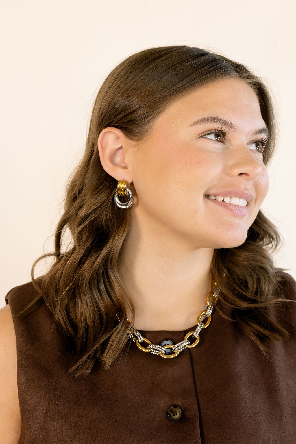 A woman with brown hair is shown in a close-up portrait, wearing a chunky gold and silver chain-link necklace and matching hoop earrings against a plain light background.