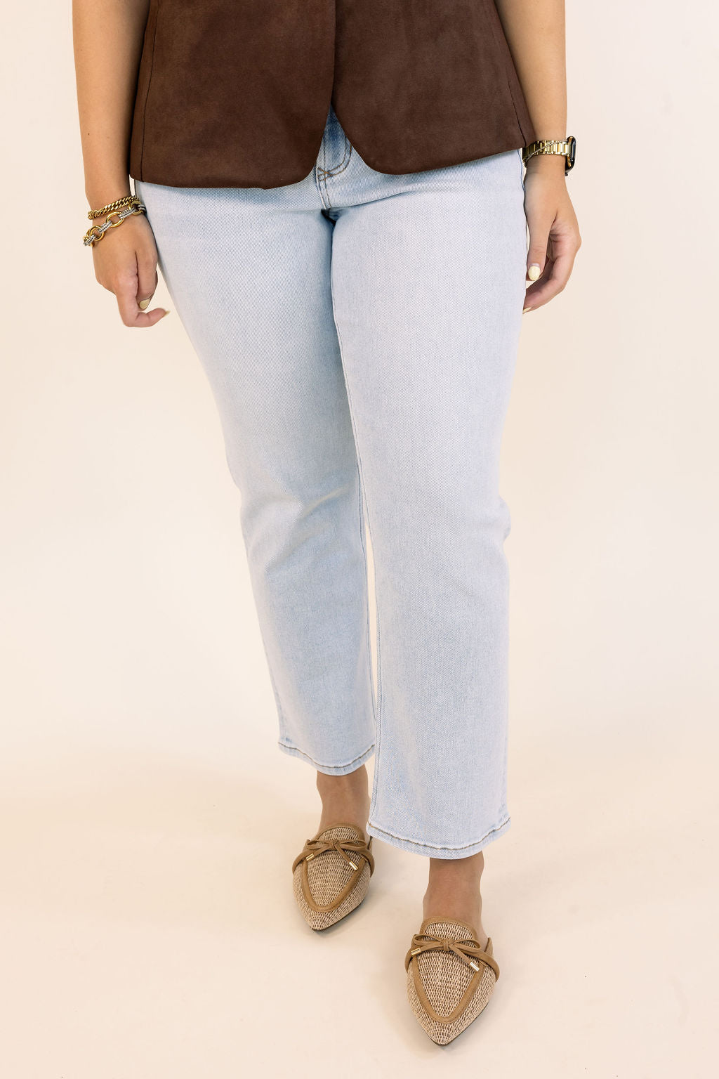 A person wearing light-wash light blue cropped jeans, a brown suede-like top, and tan woven mule flats, shown from the waist down against a plain light beige studio background.