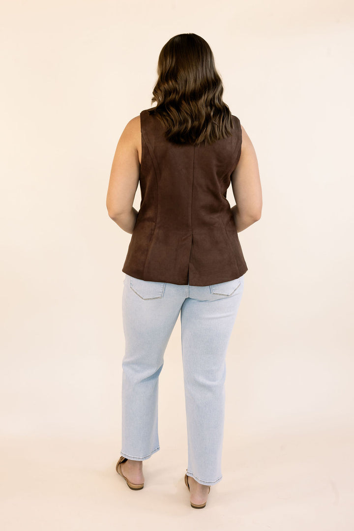A woman viewed from behind wearing a sleeveless dark brown vest and light blue jeans, standing against a plain cream studio background.