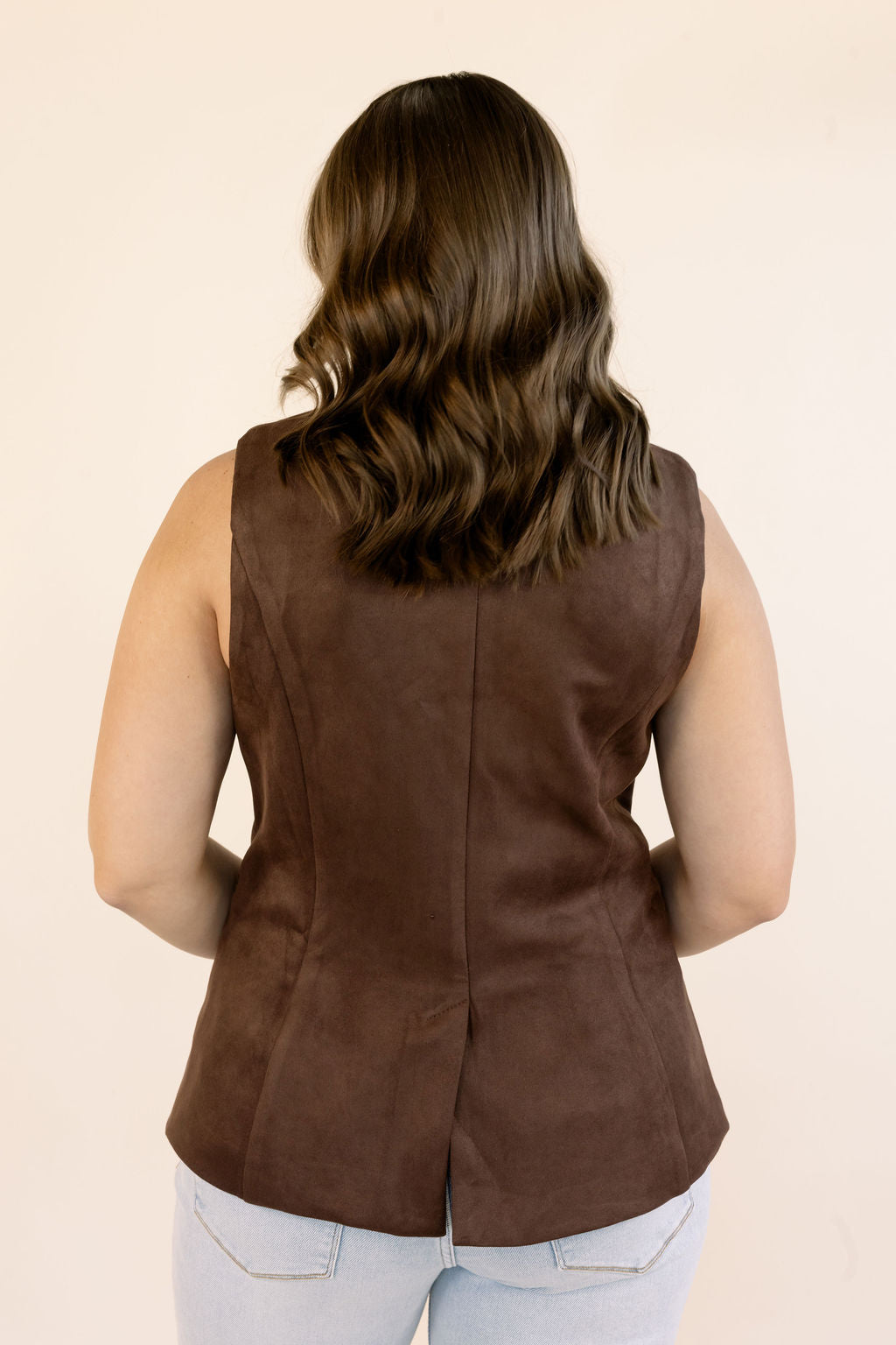 A woman viewed from the back wearing a sleeveless brown peplum-style vest with structured seams, paired with light blue jeans, against a plain beige studio background.