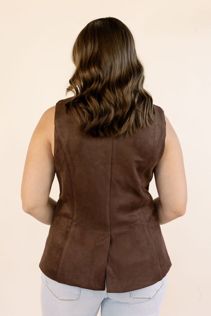 A woman viewed from the back wearing a sleeveless brown peplum-style vest with structured seams, paired with light blue jeans, against a plain beige studio background.
