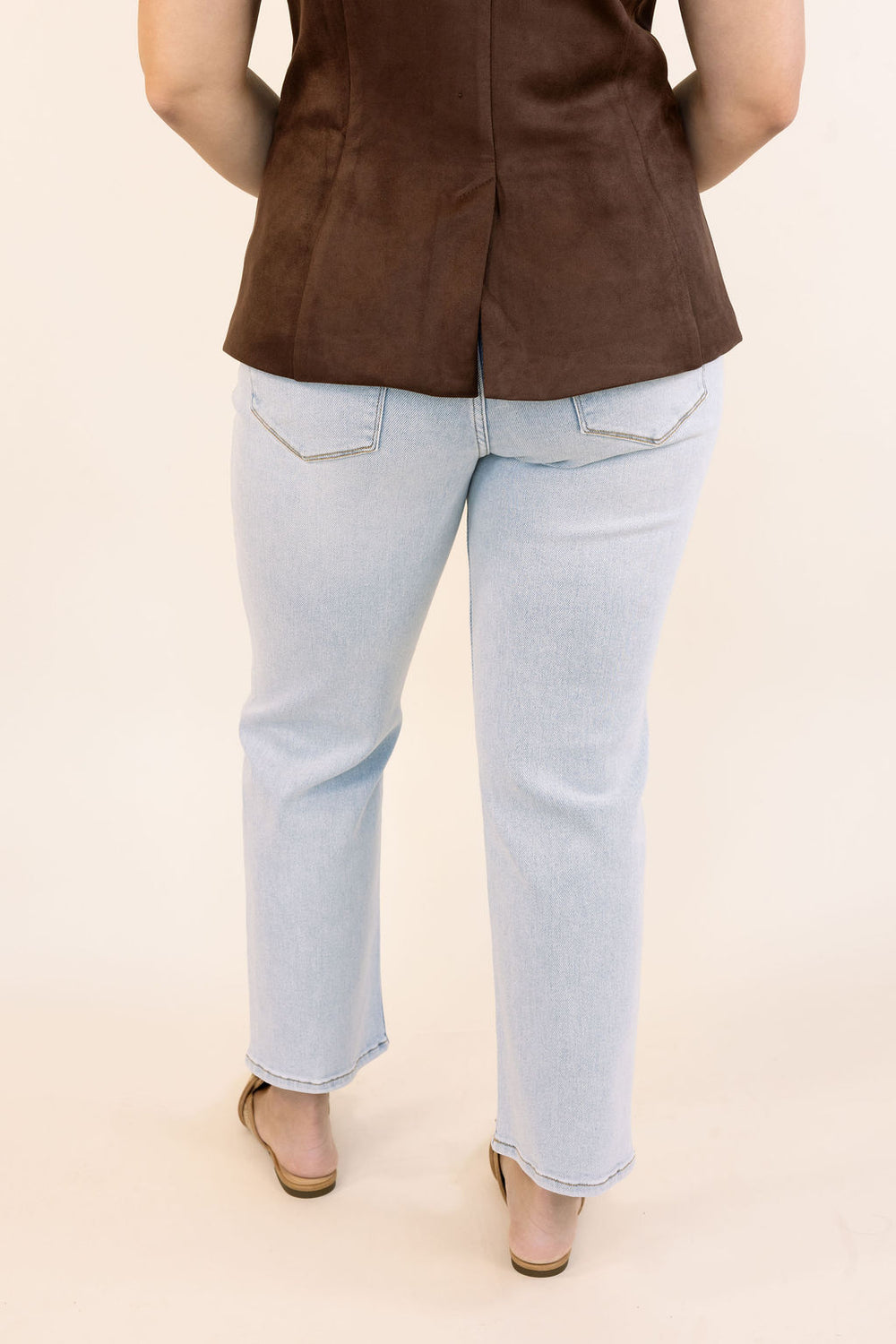 A person shown from the back wearing light-wash blue jeans, a brown sleeveless top, and tan sandals against a plain cream studio background.