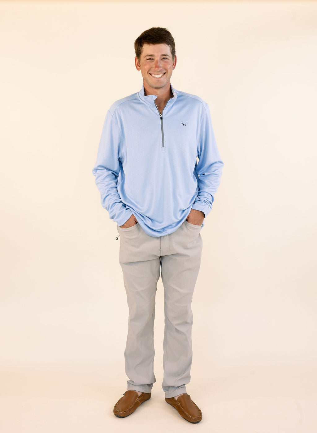 A smiling man wearing a light blue long-sleeve quarter-zip pullover, khaki pants, and brown loafers, standing against a solid cream-colored background.