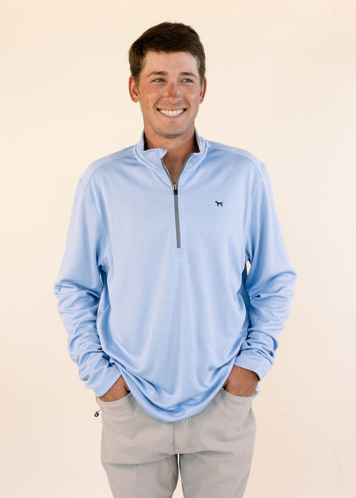 A smiling man wearing a light blue long-sleeve quarter-zip pullover with a small black dog logo on the left chest, paired with khaki pants, against a plain beige background.
