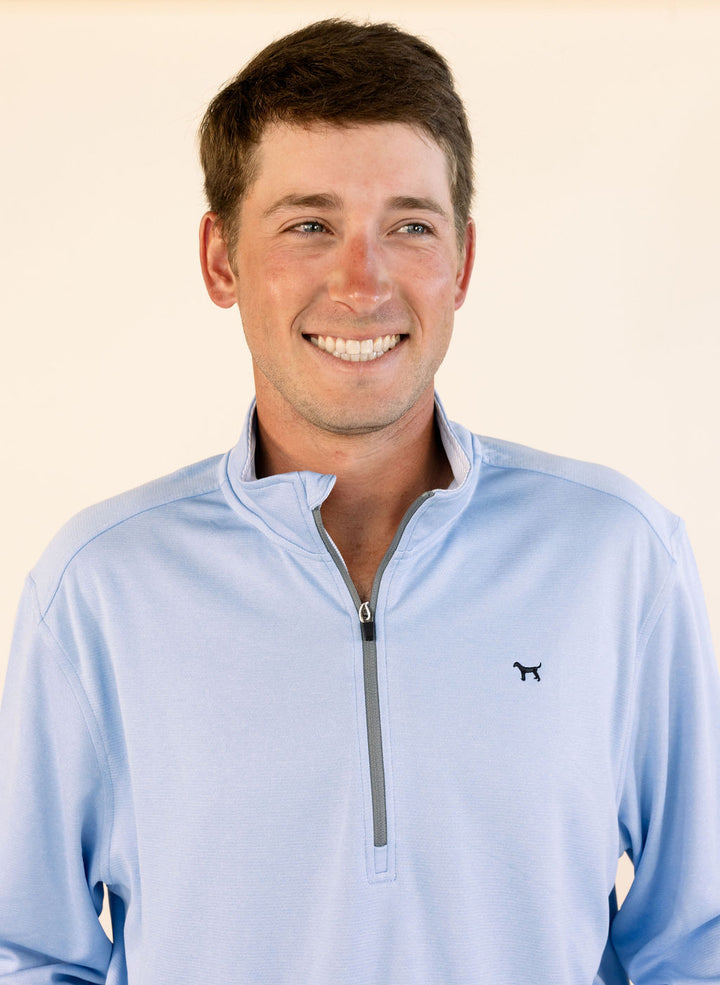 A smiling man wearing a light blue quarter-zip pullover with a small black dog logo on the left chest, photographed against a plain light beige background.