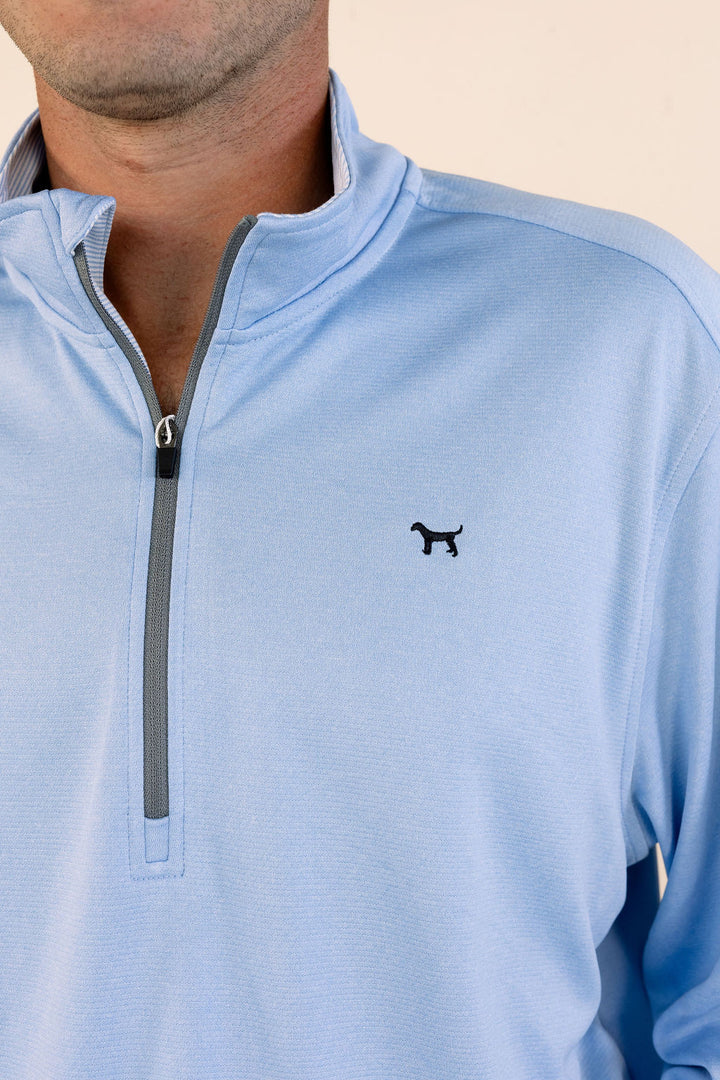A close-up of a man wearing a light blue quarter-zip pullover with a gray zipper track and a small black dog logo on the left chest.