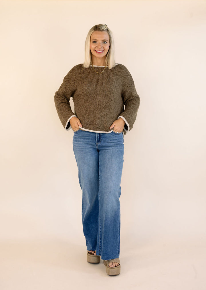 A woman modeling a mocha knit sweater with white cuffs and hem, paired with blue jeans and beige platform sandals, standing against a plain cream background.