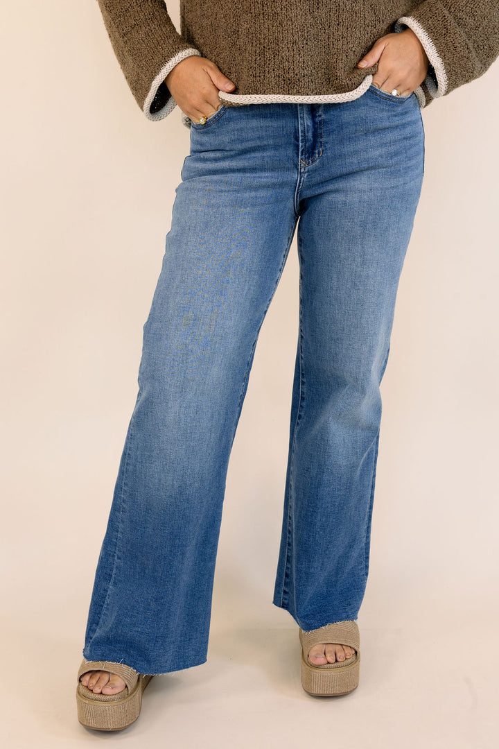 A person wearing medium-wash blue flare jeans with a brown knit sweater and beige platform sandals, standing against a plain light beige studio background.