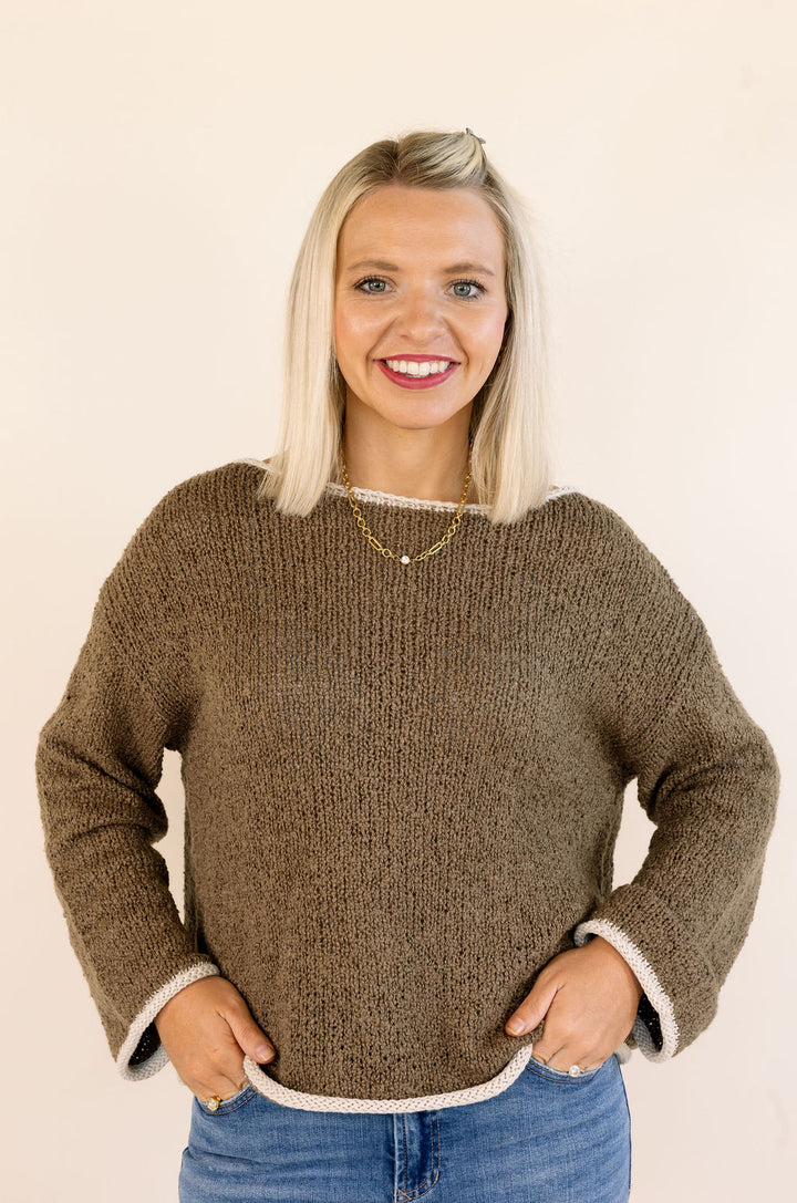 A smiling blonde woman wearing a mocha textured knit sweater with white ribbed trim on the cuffs and hem, paired with blue jeans, against a plain cream background.