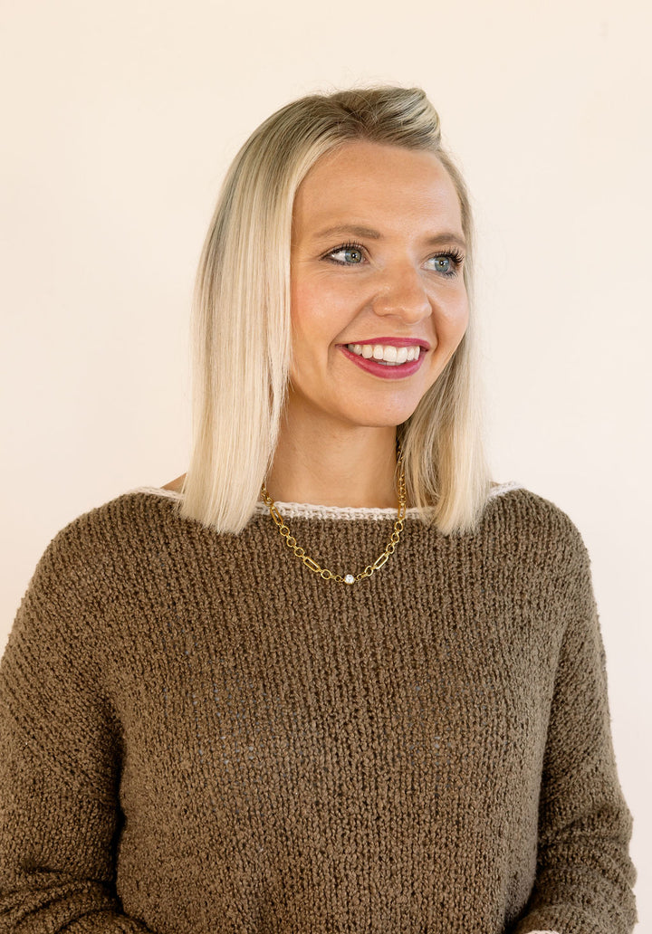 A smiling blonde woman wearing a mocha brown chunky knit sweater and a delicate gold necklace, photographed against a plain off-white background.