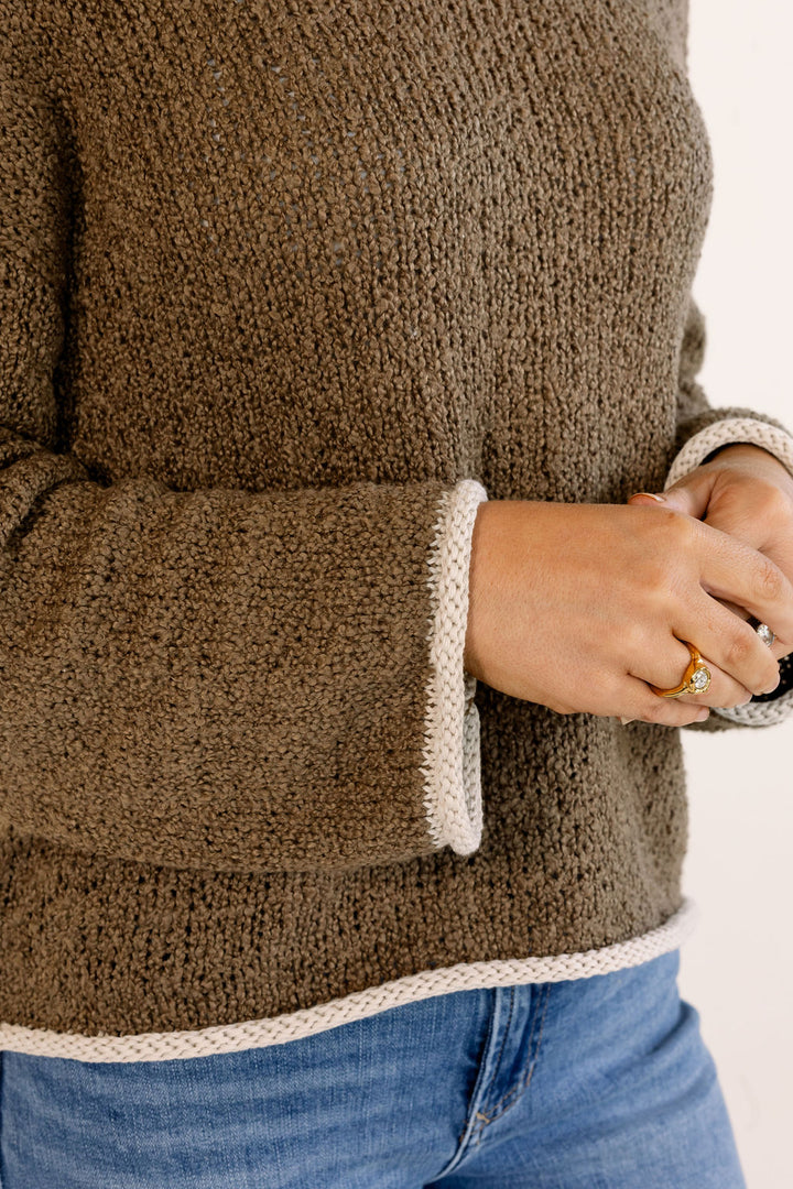 A close-up of a person wearing a chunky knit brown sweater with cream ribbed trim on the cuffs and hem, paired with blue jeans.