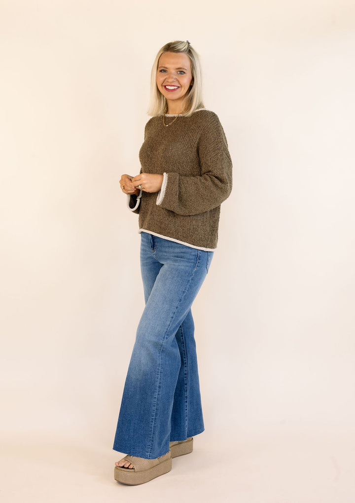 A woman modeling a mocha brown knit sweater with white cuffs and hem, paired with blue flare jeans and beige platform sandals, standing against a plain cream background.