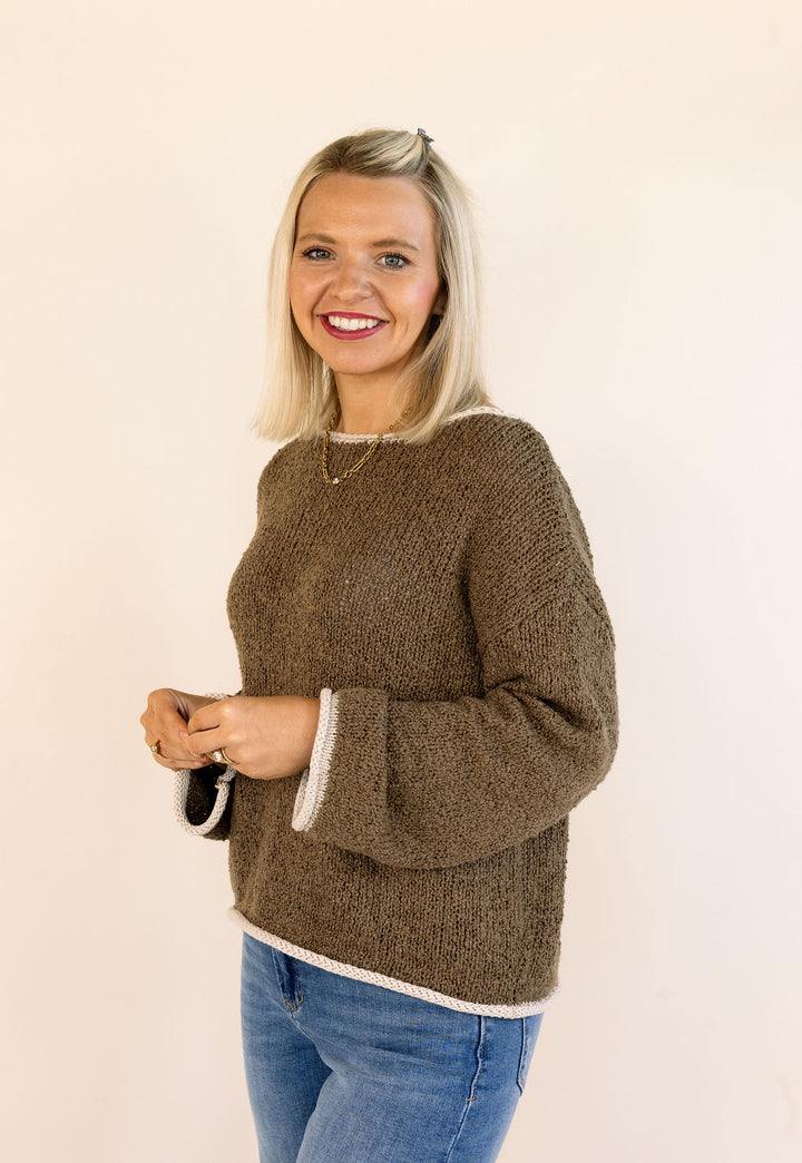 A smiling blonde woman wearing a mocha brown textured knit sweater with white ribbed trim on the cuffs and hem, paired with blue jeans, standing against a plain off-white background.