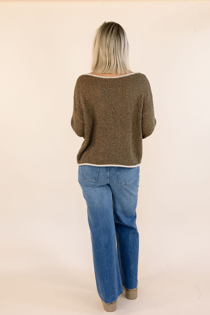 A woman viewed from behind wearing a mocha brown textured knit sweater and blue jeans, standing against a plain beige studio background.