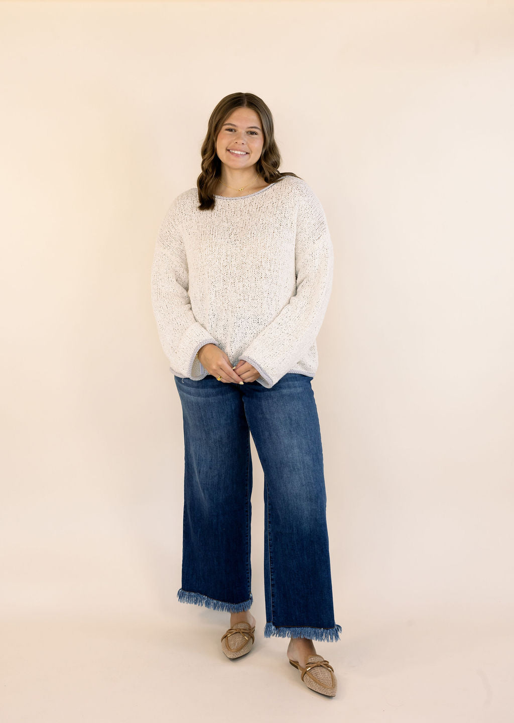 A woman modeling an oatmeal colored, textured knit sweater and wide-leg blue jeans with frayed hems, standing against a solid light beige studio background.