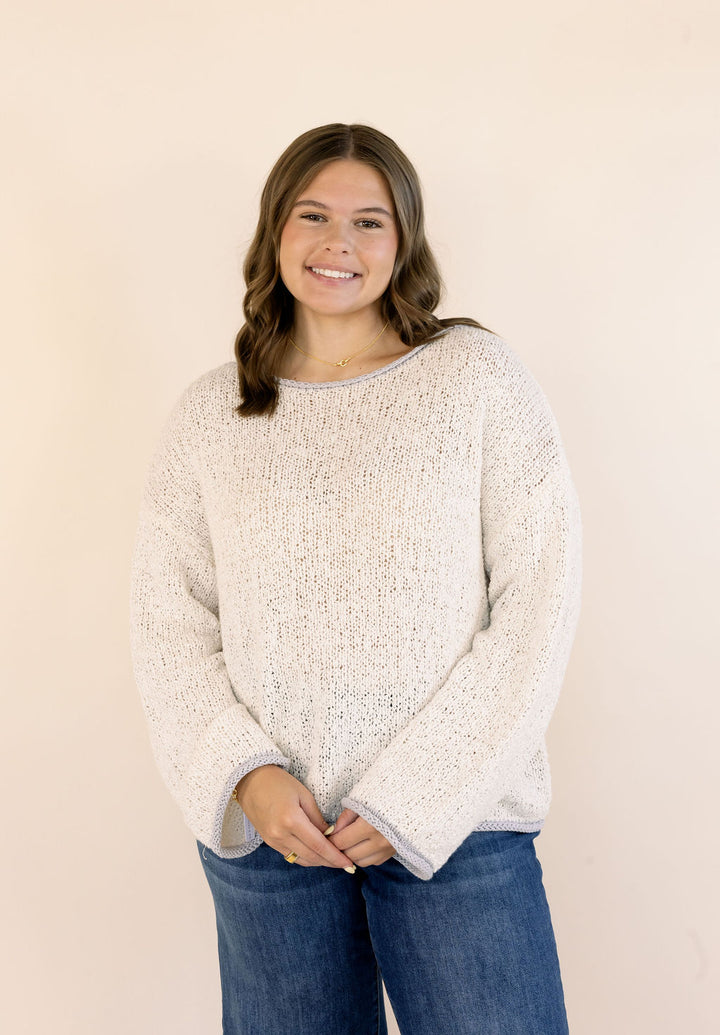 A woman wearing a oatmeal-colored, textured knit crewneck sweater and blue jeans, standing against a solid light beige background.