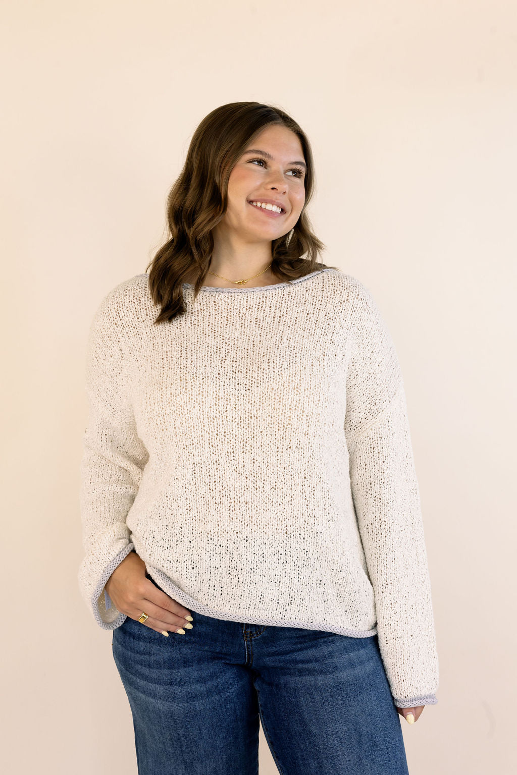 A woman wearing an oatmeal-colored, textured knit sweater and blue jeans, standing against a solid light beige background.
