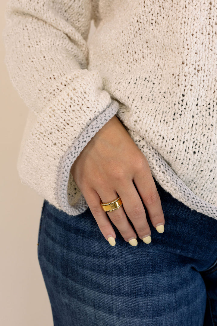 A close-up of a person wearing a chunky gold ring on their finger, styled with a cream-colored knit sweater and blue jeans.
