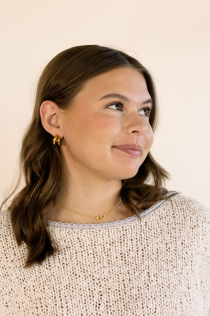 A woman with brown hair wearing a cream knit sweater, a delicate gold necklace, and a small gold double hoop earring, looking to the side against a plain light beige background.