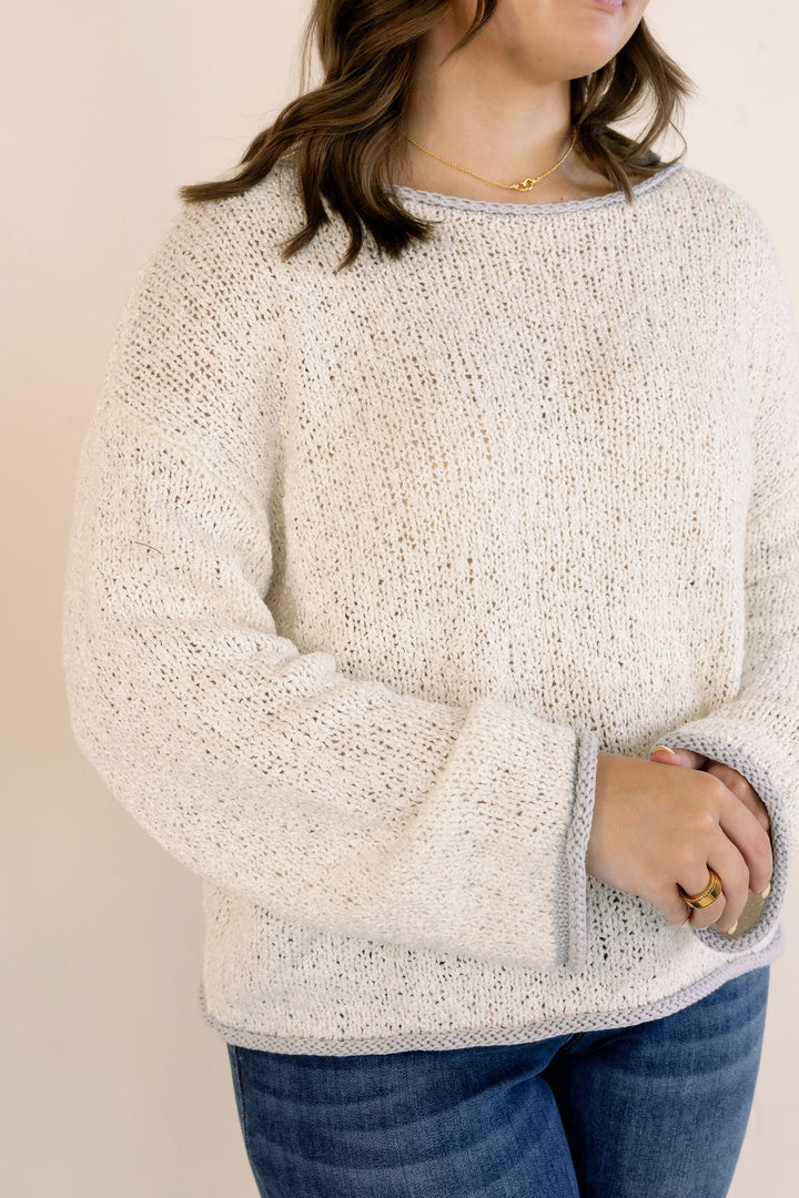 A woman wearing an oatmeal-colored, textured knit sweater with blue jeans, shown from the neck down against a plain beige background.