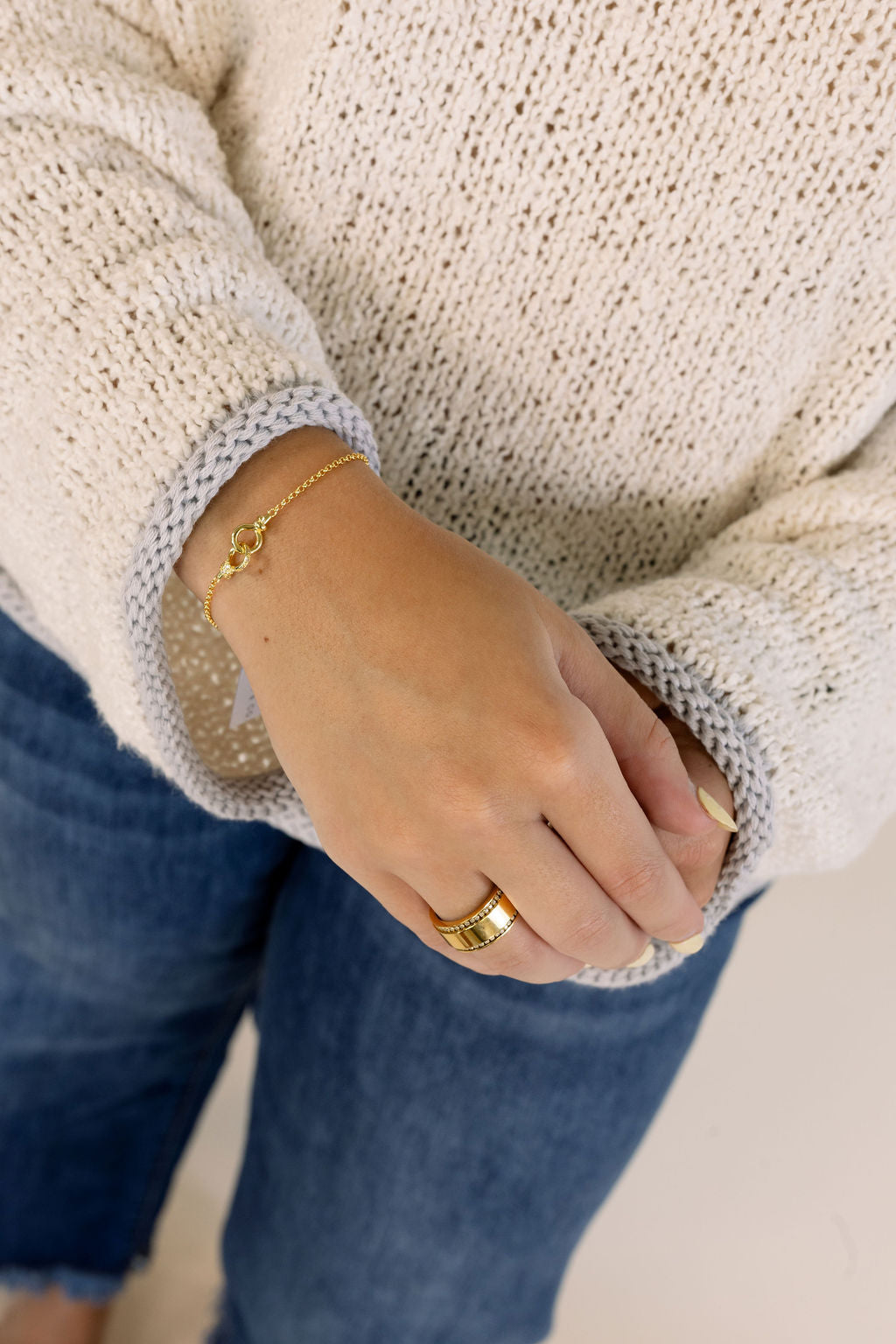 A close-up lifestyle shot of a person wearing a chunky knit cream sweater, blue jeans, a delicate gold chain bracelet with interlocking charms, and a wide gold ring.