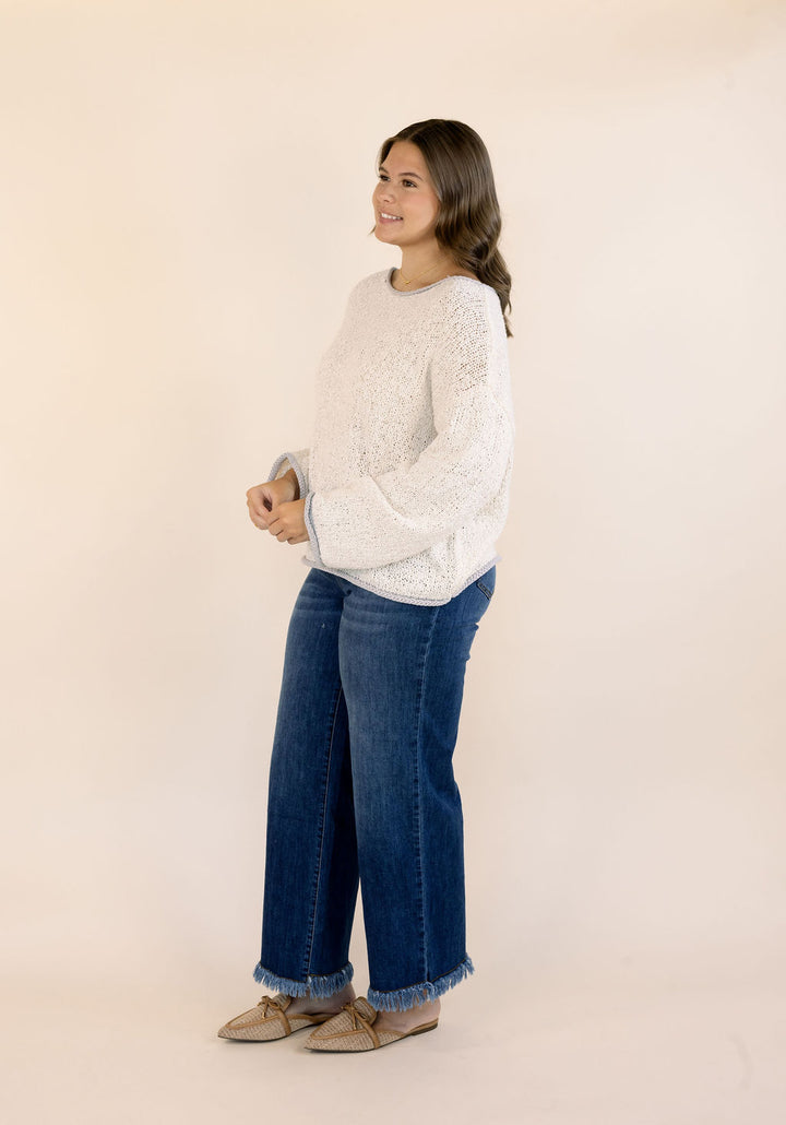 A woman modeling an oatmeal textured knit sweater and blue wide-leg jeans with frayed hems, paired with tan loafers, against a solid light beige studio background.