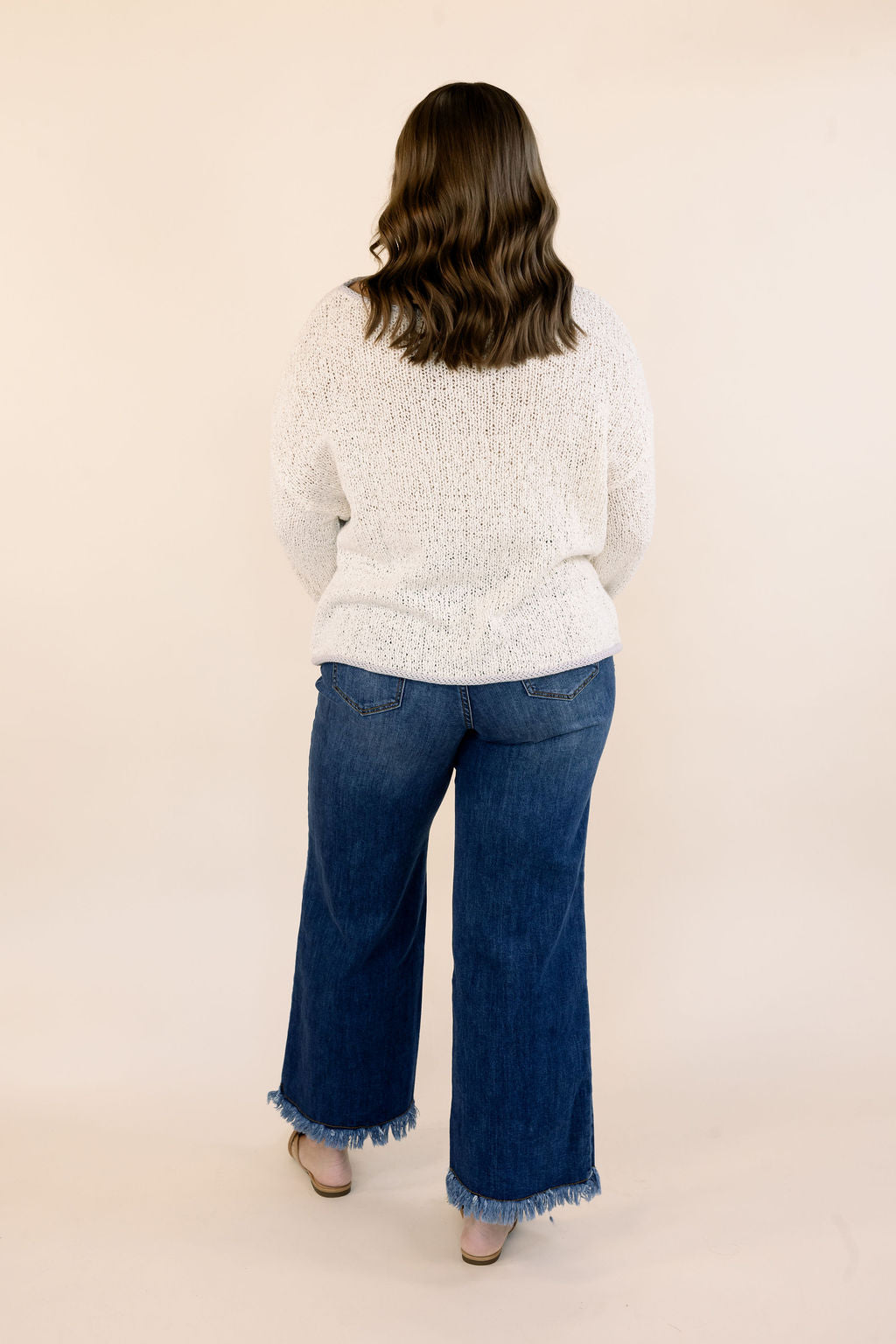 A woman viewed from behind wearing an oatmeal textured knit sweater and blue wide-leg jeans with frayed hems, standing against a plain beige studio background.