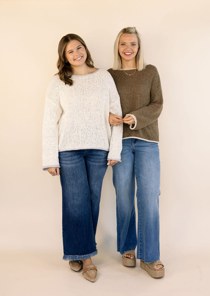 Two women standing side-by-side wearing knit sweaters and wide-leg jeans against a solid beige studio background.