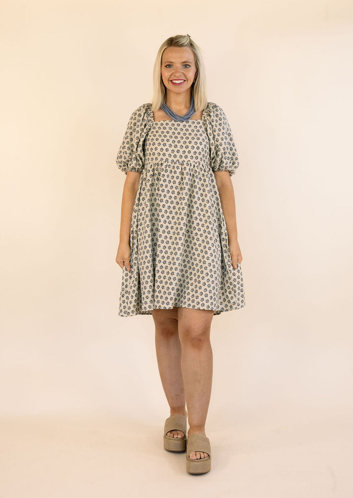 A woman modeling a short puff-sleeve dress with a brown and blue floral pattern, paired with beige platform sandals against a plain beige studio background.