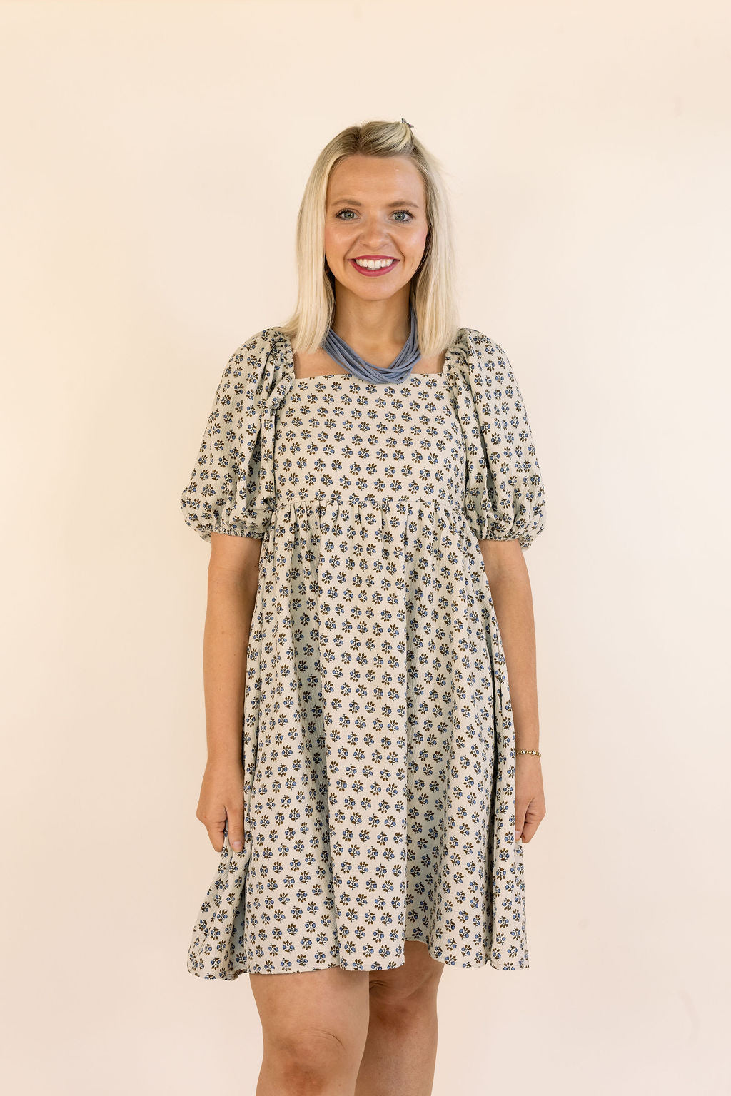 A smiling blonde woman wearing a short puff-sleeve dress with a blue and brown floral pattern, standing against a plain beige background.