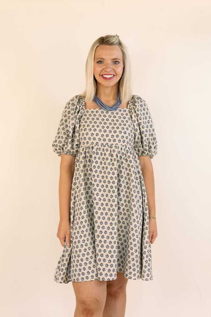 A smiling blonde woman wearing a short puff-sleeve dress with a blue and brown floral pattern, standing against a plain beige background.