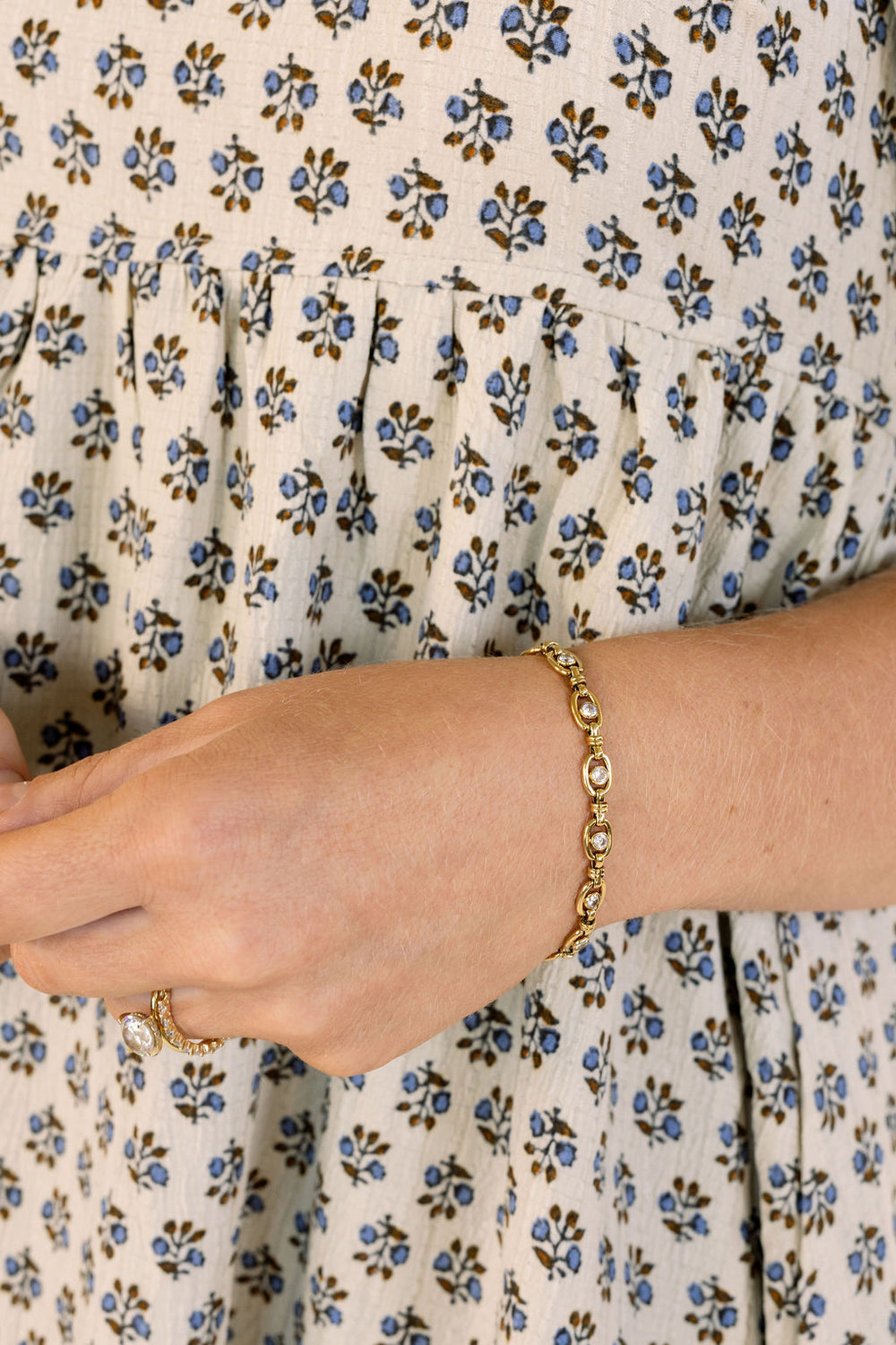 A gold-toned chain bracelet with clear oval stones worn on a person's wrist, shown against a cream dress with a blue and brown floral print.
