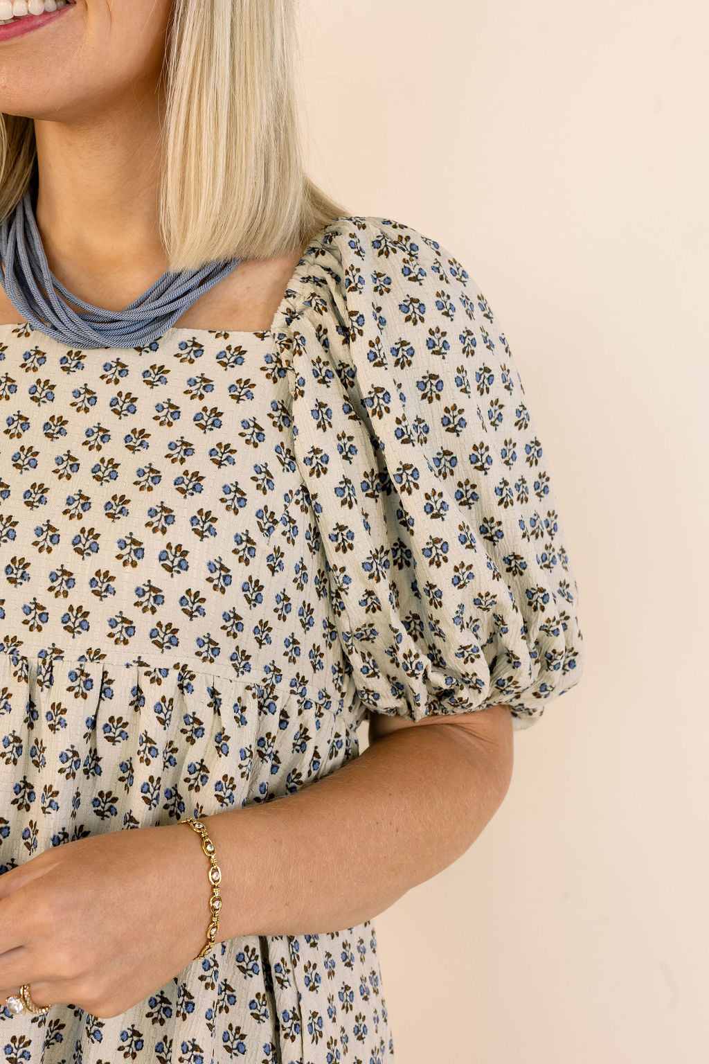 A close-up of a woman wearing a cream-colored puff-sleeve dress with a small blue and black floral pattern, styled with a layered blue necklace and a gold bracelet against a solid beige background.