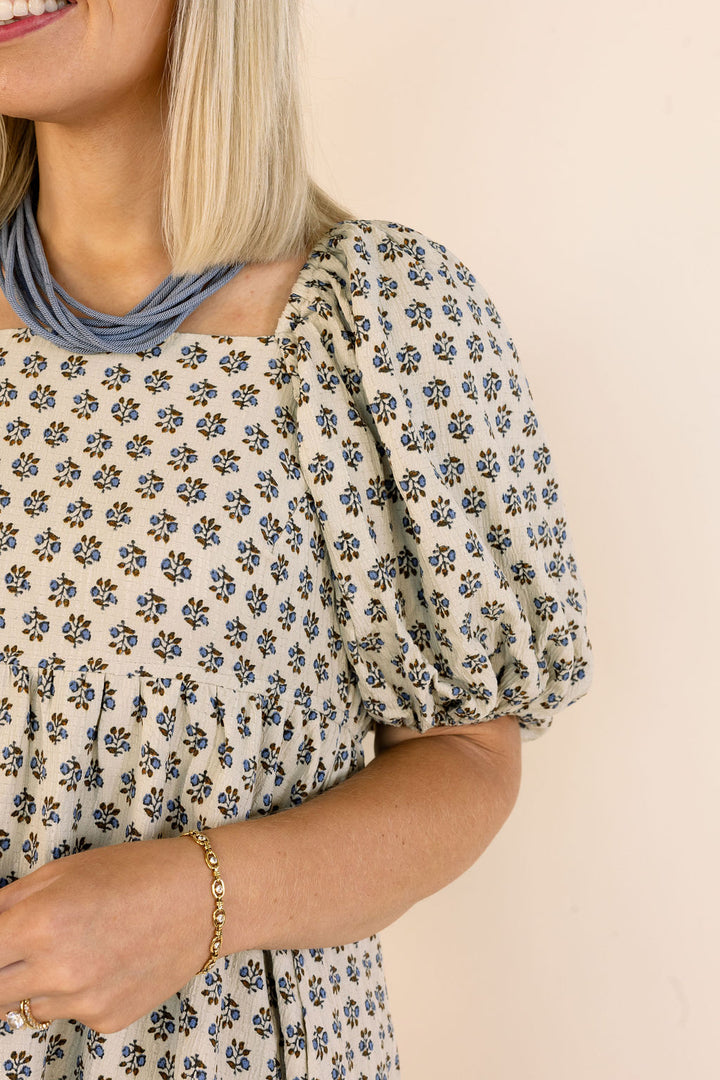 A close-up of a woman wearing a cream-colored puff-sleeve dress with a small blue and black floral pattern, styled with a layered blue necklace and a gold bracelet against a solid beige background.