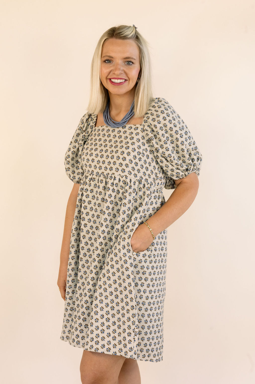 A woman wearing a short puff-sleeve dress with an all-over small floral print in blue and brown, standing against a plain light beige background.