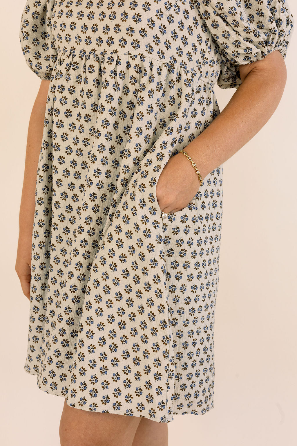 A close-up of a person wearing a short puff-sleeve dress with an all-over small floral print in blue and brown on a warm cream background. The dress has side pockets, and the model&