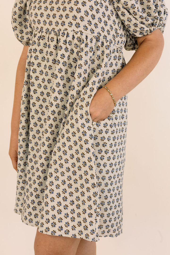 A close-up of a person wearing a short puff-sleeve dress with an all-over small floral print in blue and brown on a warm cream background. The dress has side pockets, and the model&